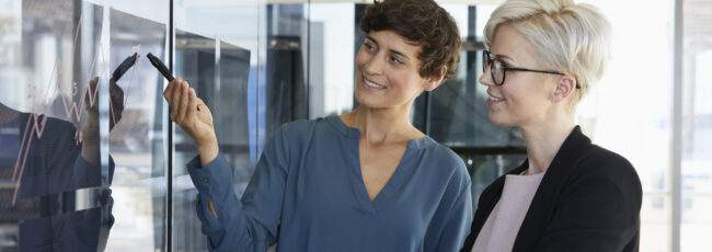 Two smiling businesswomen looking at chart on glass pane in office